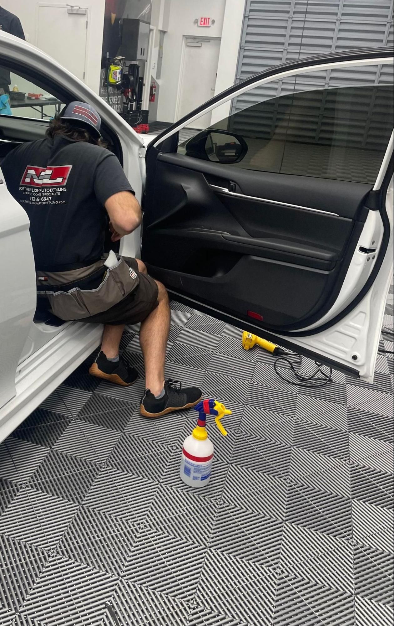 Person applying tint to a car window inside a shop. They're seated with tools and a spray bottle nearby.