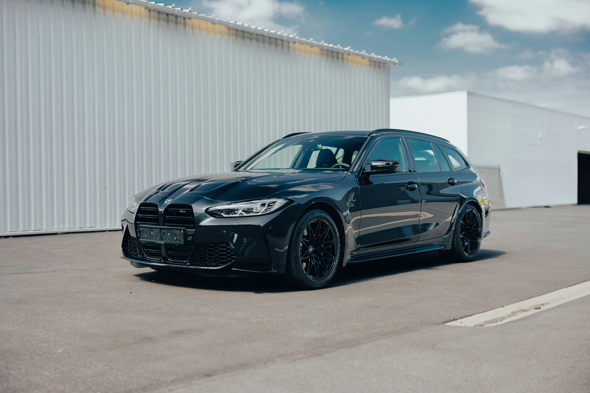 Black BMW M3 Touring parked on asphalt in front of a white building.