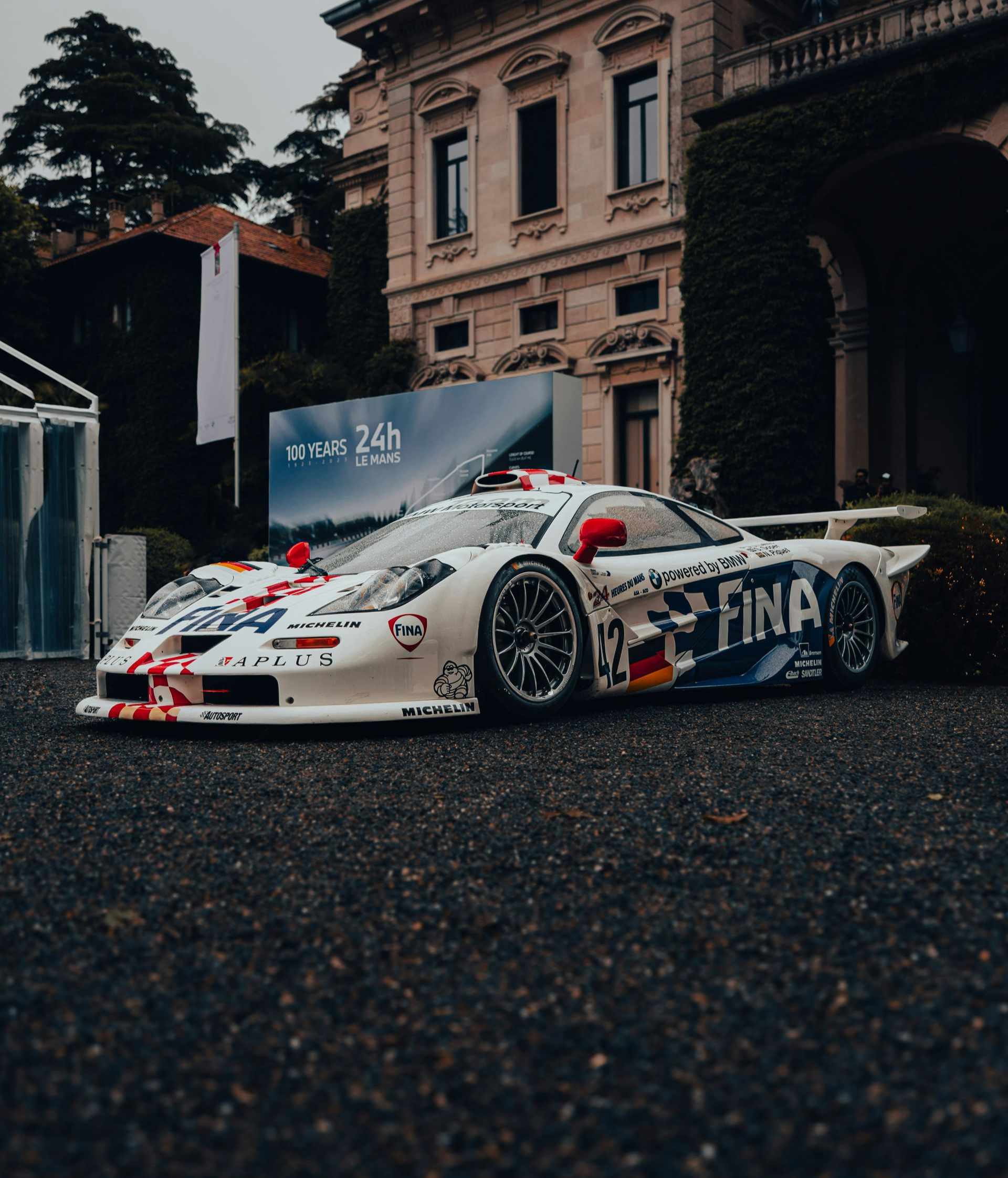 White race car with colorful livery, parked on cobblestones in front of a grand building.