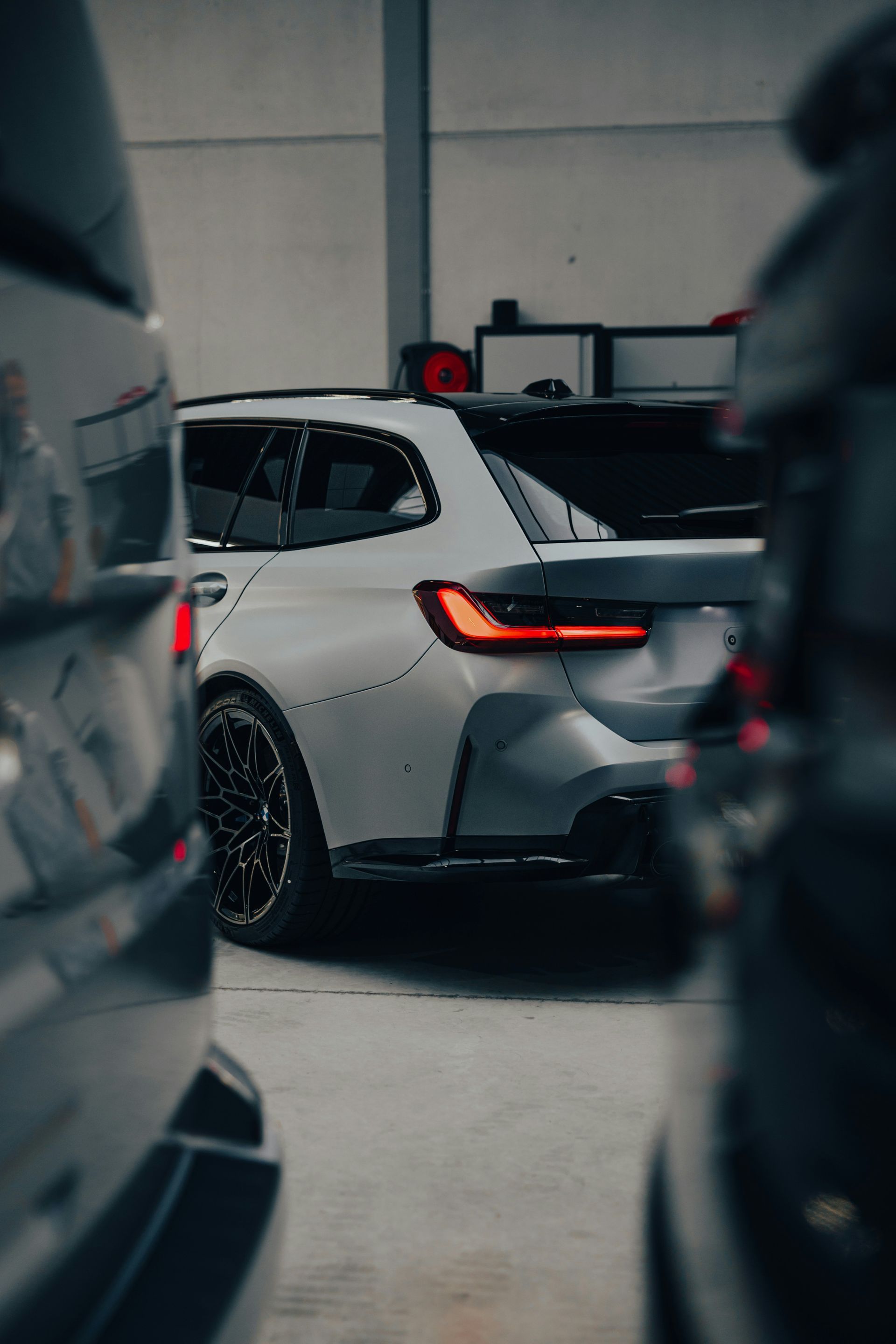 Gray BMW station wagon in a garage, black wheels, red taillights, between two other cars.