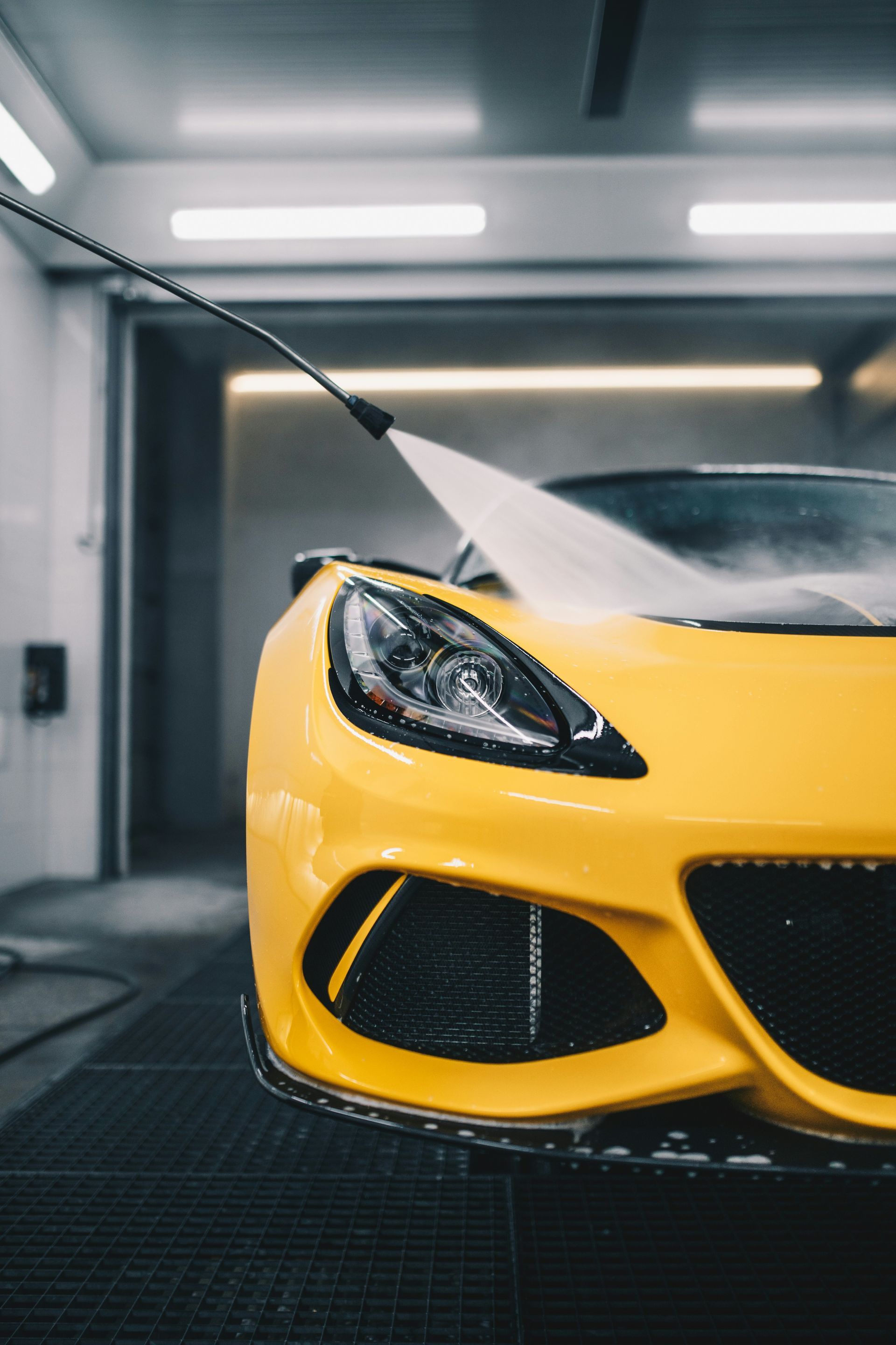 Yellow sports car being washed with a pressure washer in a car wash bay.