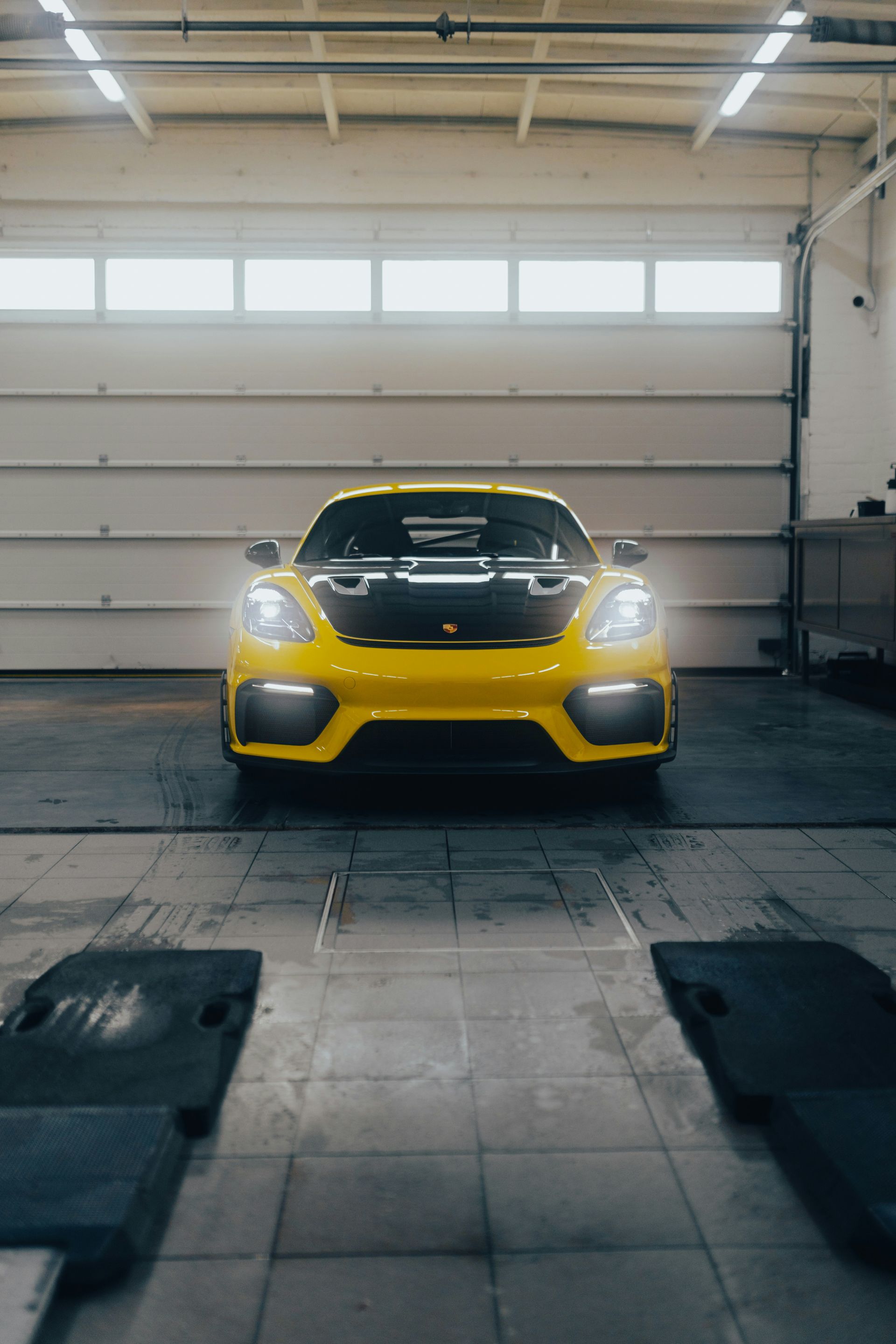 Yellow Porsche sports car in a garage with overhead door open, headlights on.
