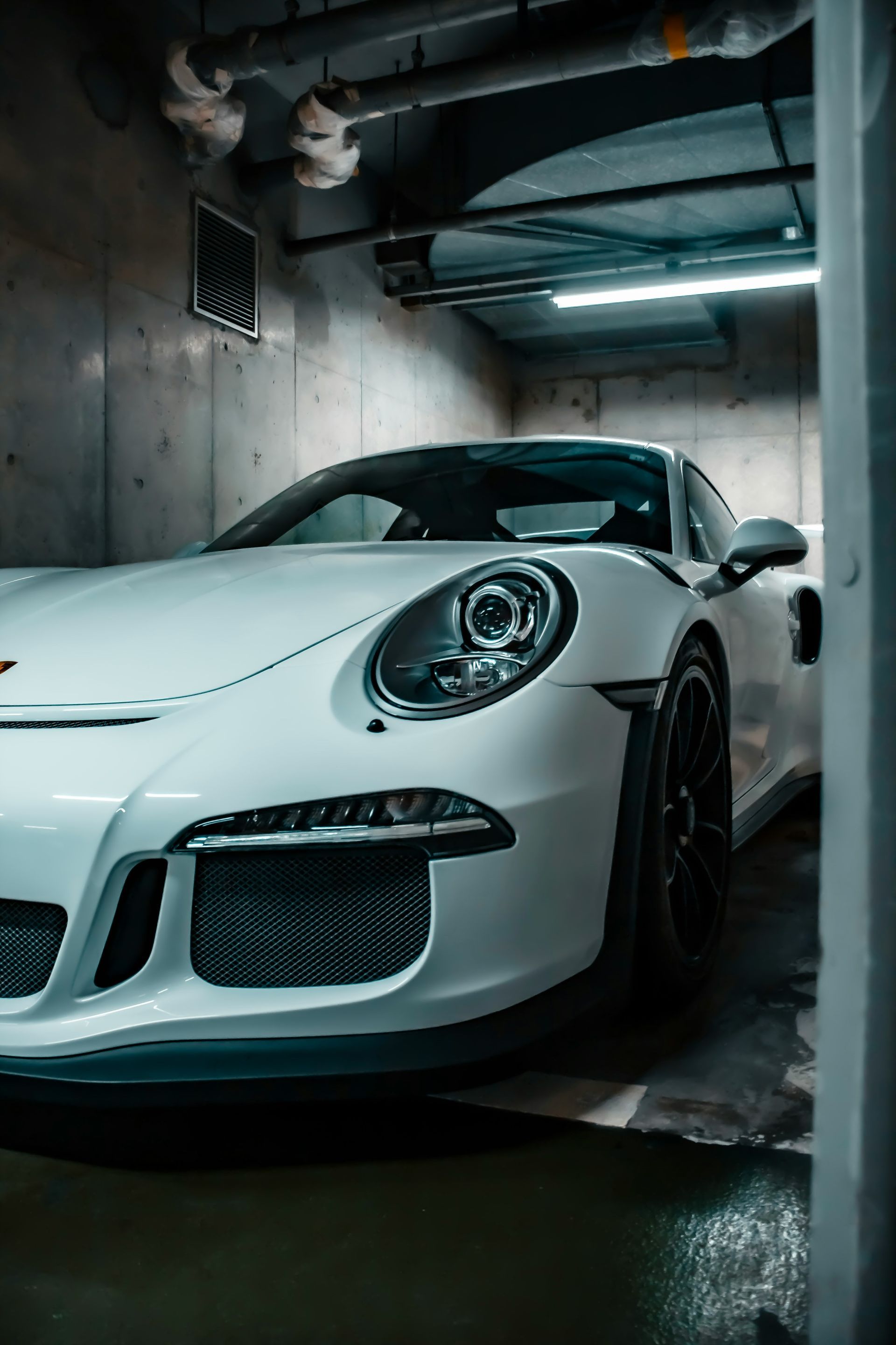 White Porsche sports car parked in a garage with concrete walls and overhead lighting.