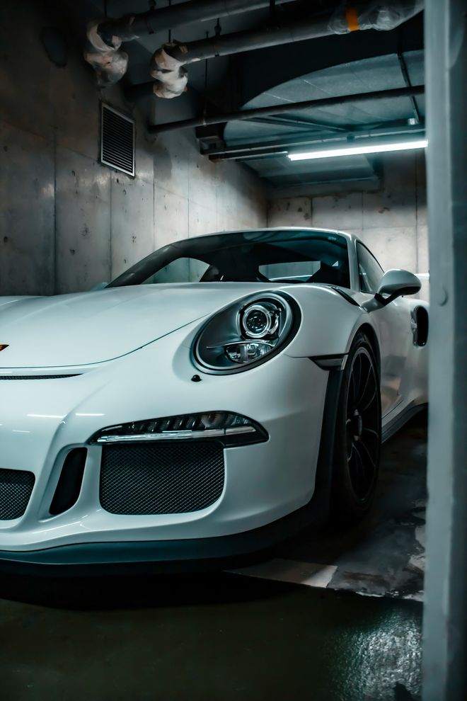 White Porsche sports car parked in a garage with concrete walls and overhead lighting.