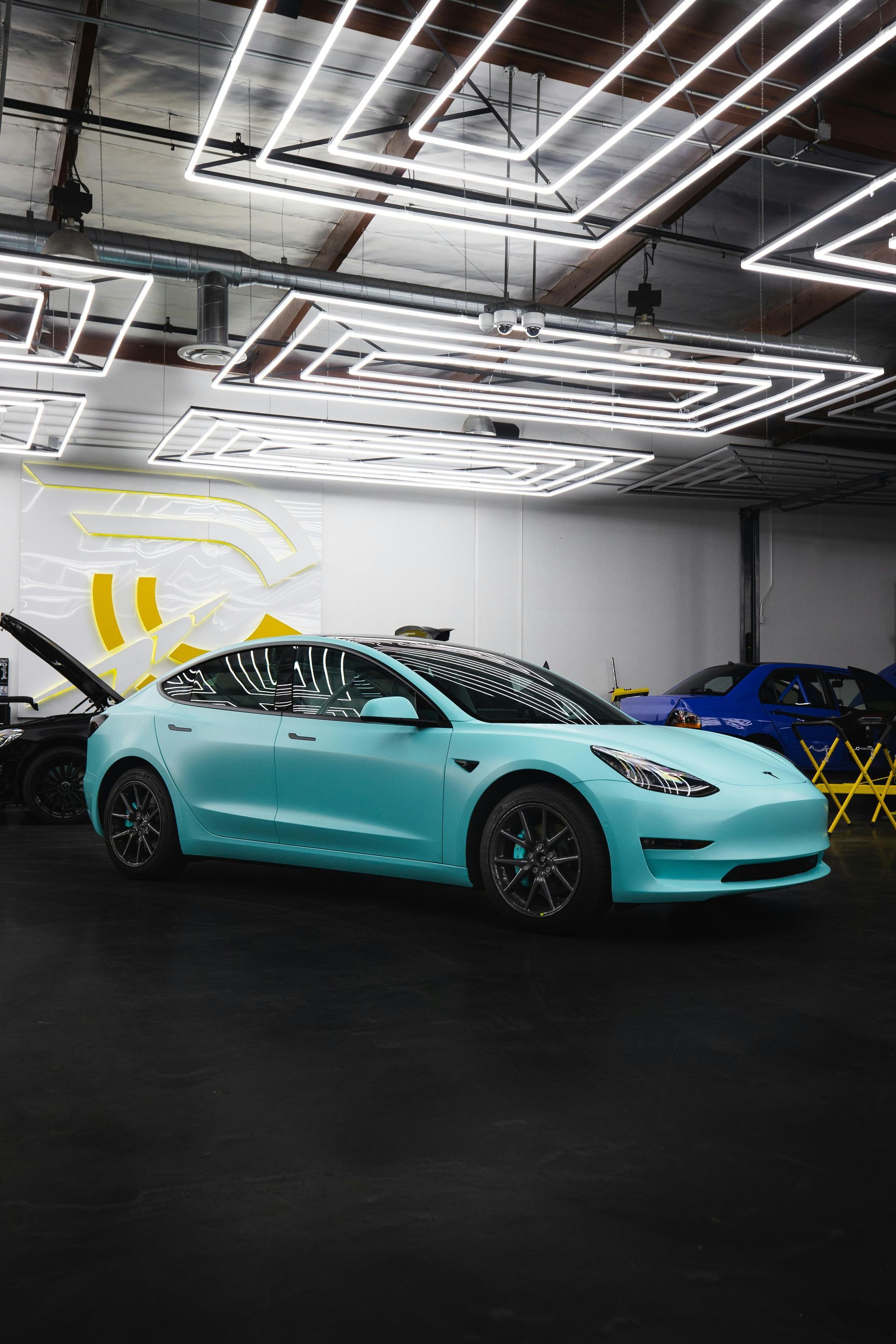 Light blue Tesla in a garage with overhead neon lights.