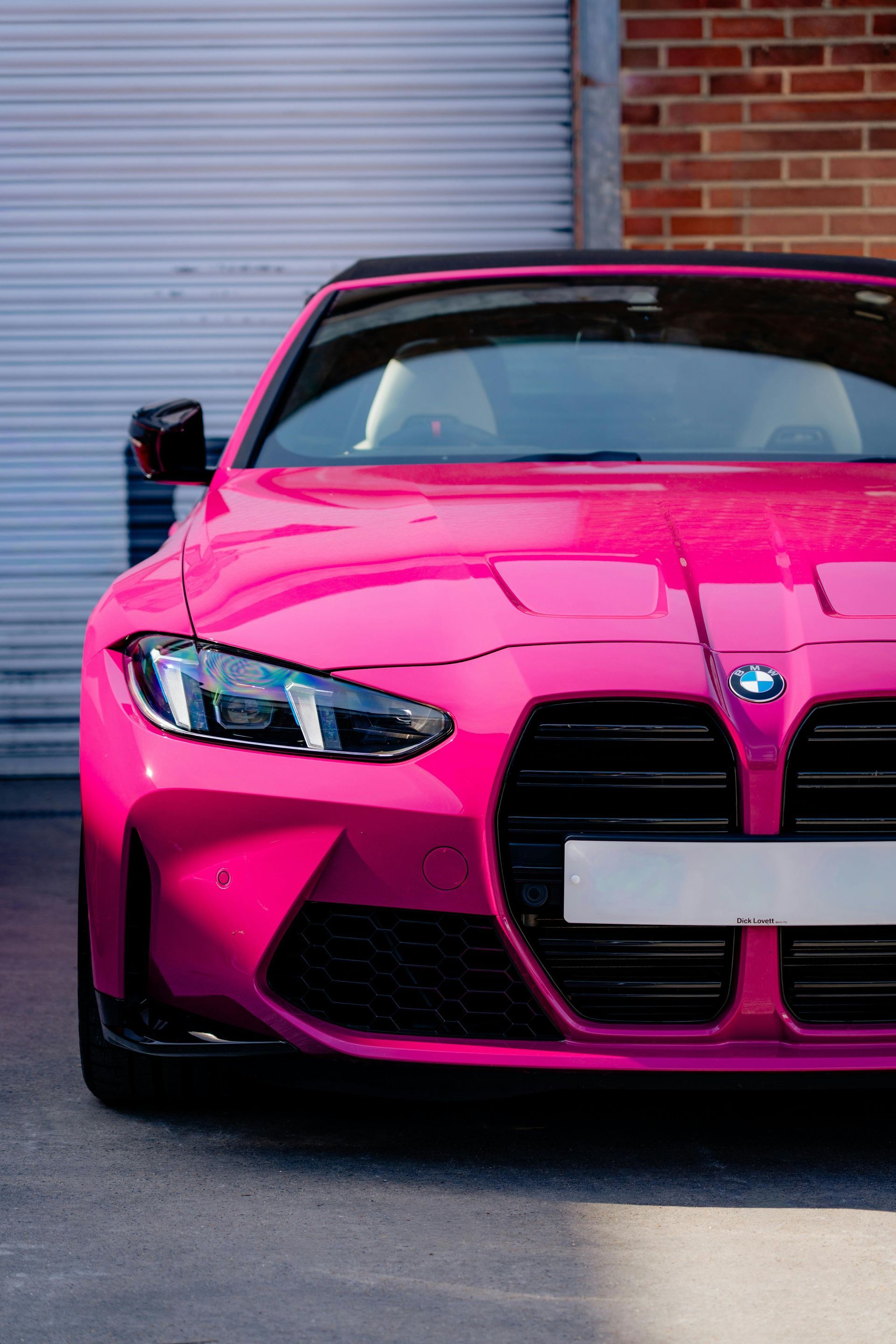 Bright pink BMW M4 sports car parked in front of a gray garage door and brick wall.