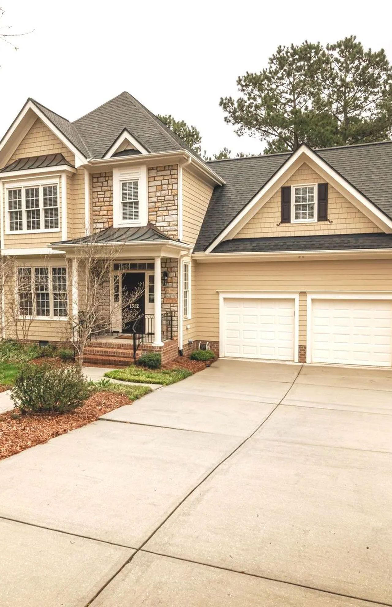 Two-story tan house with stone accents, two-car garage, and concrete driveway on an overcast day.
