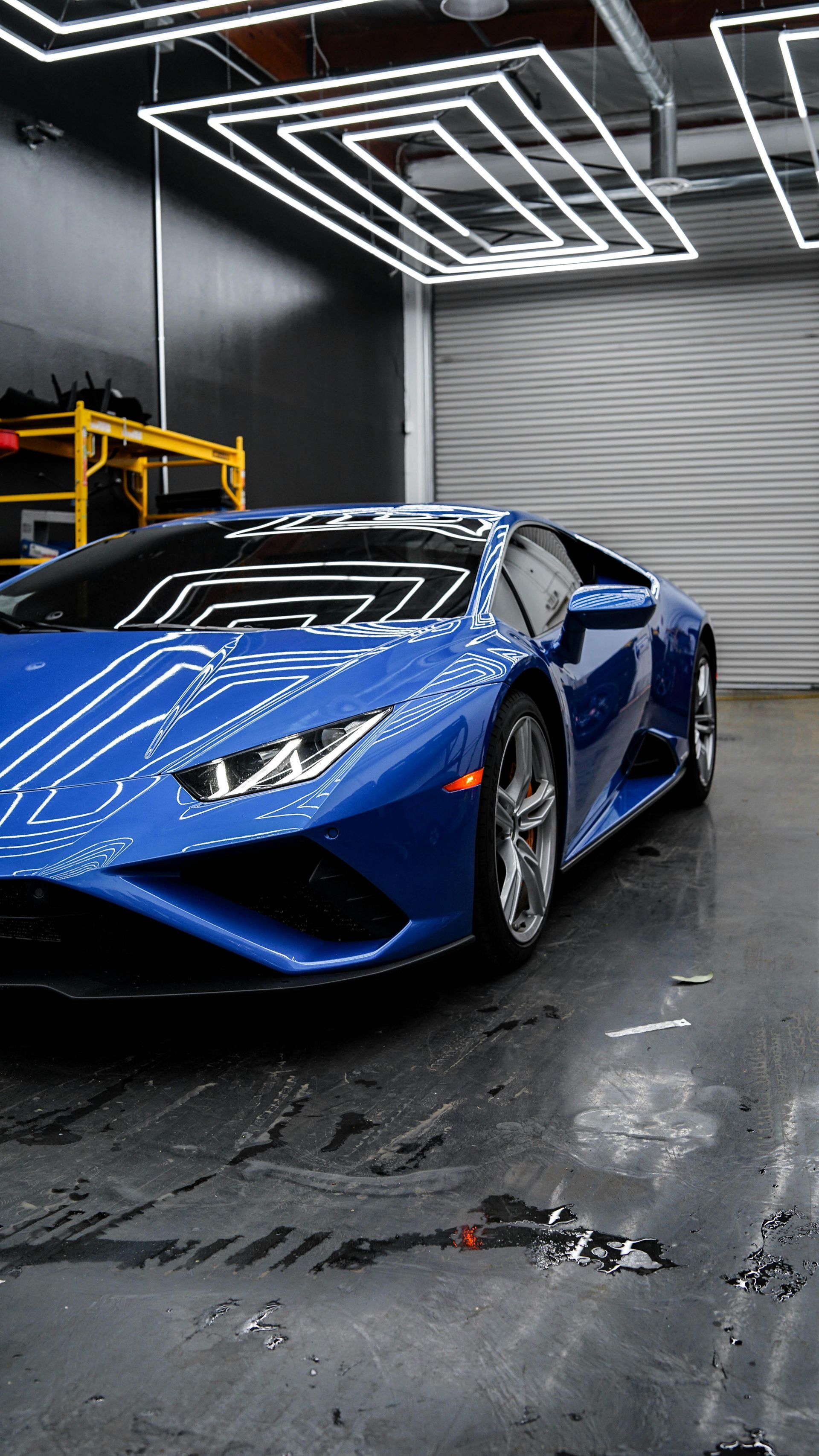Blue Lamborghini sports car parked in a garage with bright overhead lights reflecting on the shiny surface.