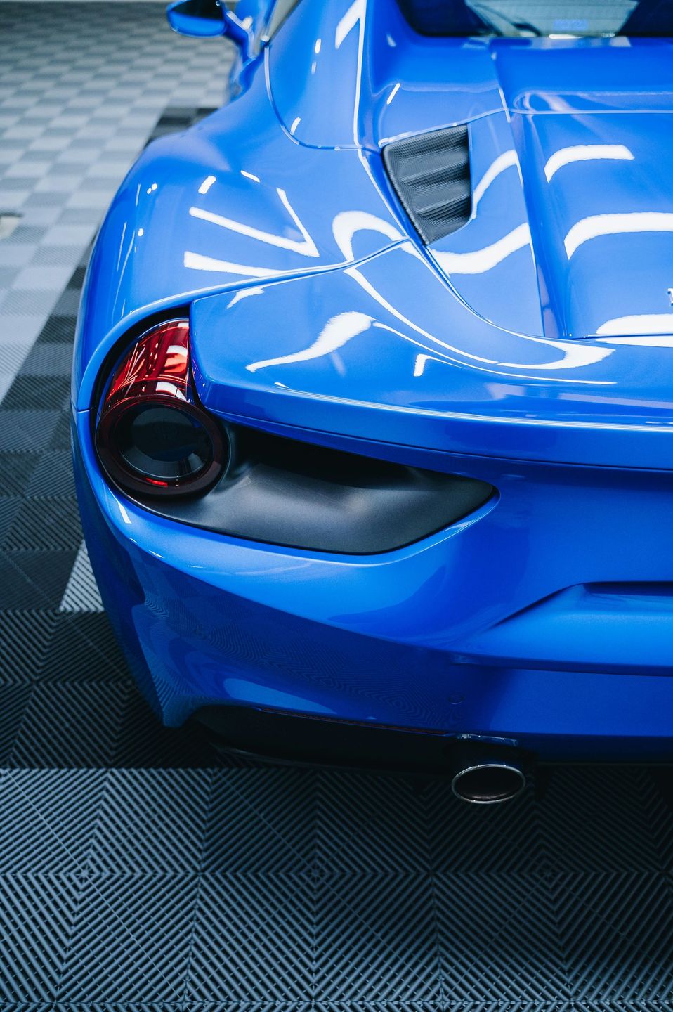 Blue sports car rear with tail light and exhaust, parked on a checkered floor.