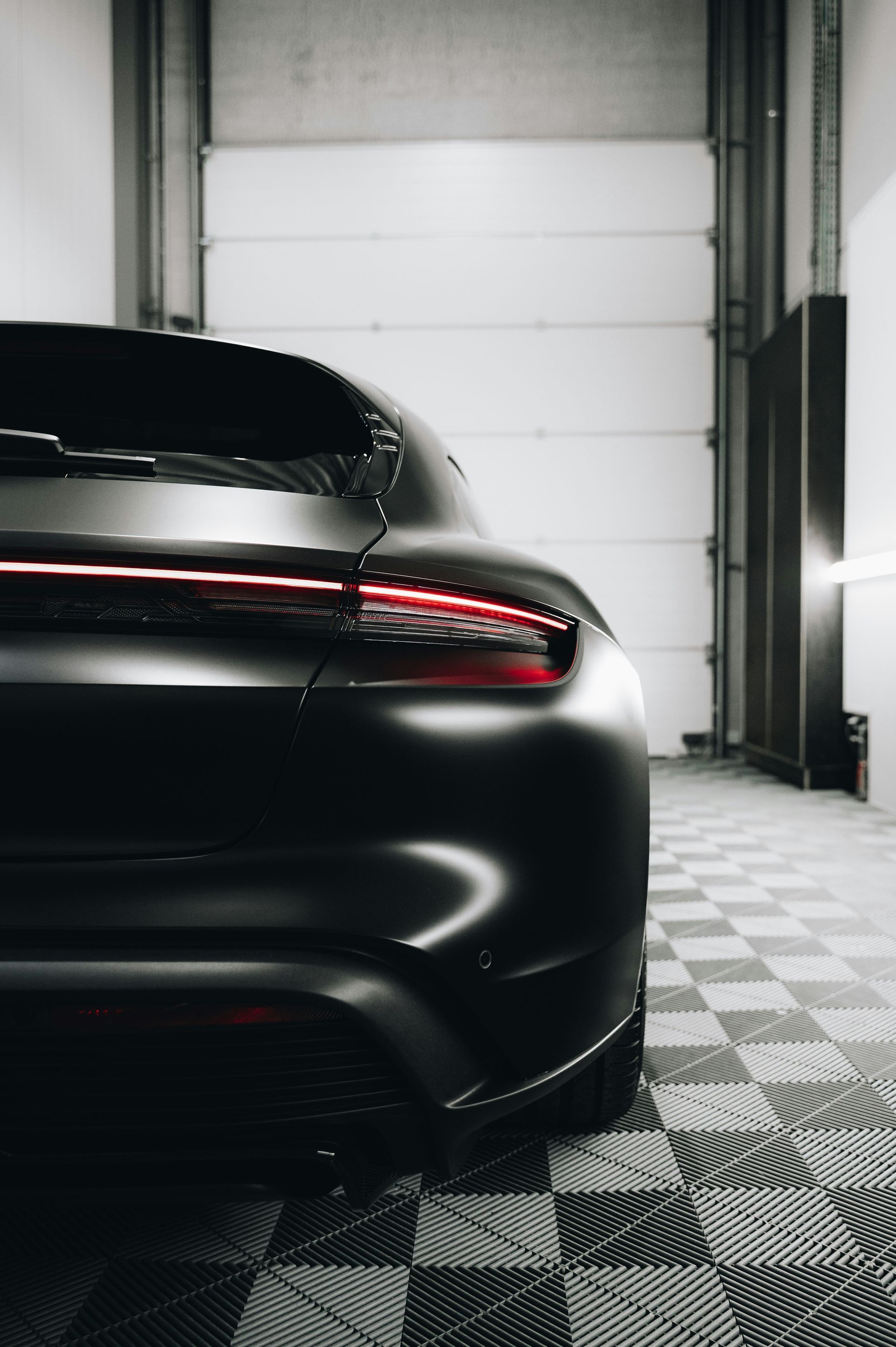 Black car parked in a garage with checkered floor; taillight illuminated.