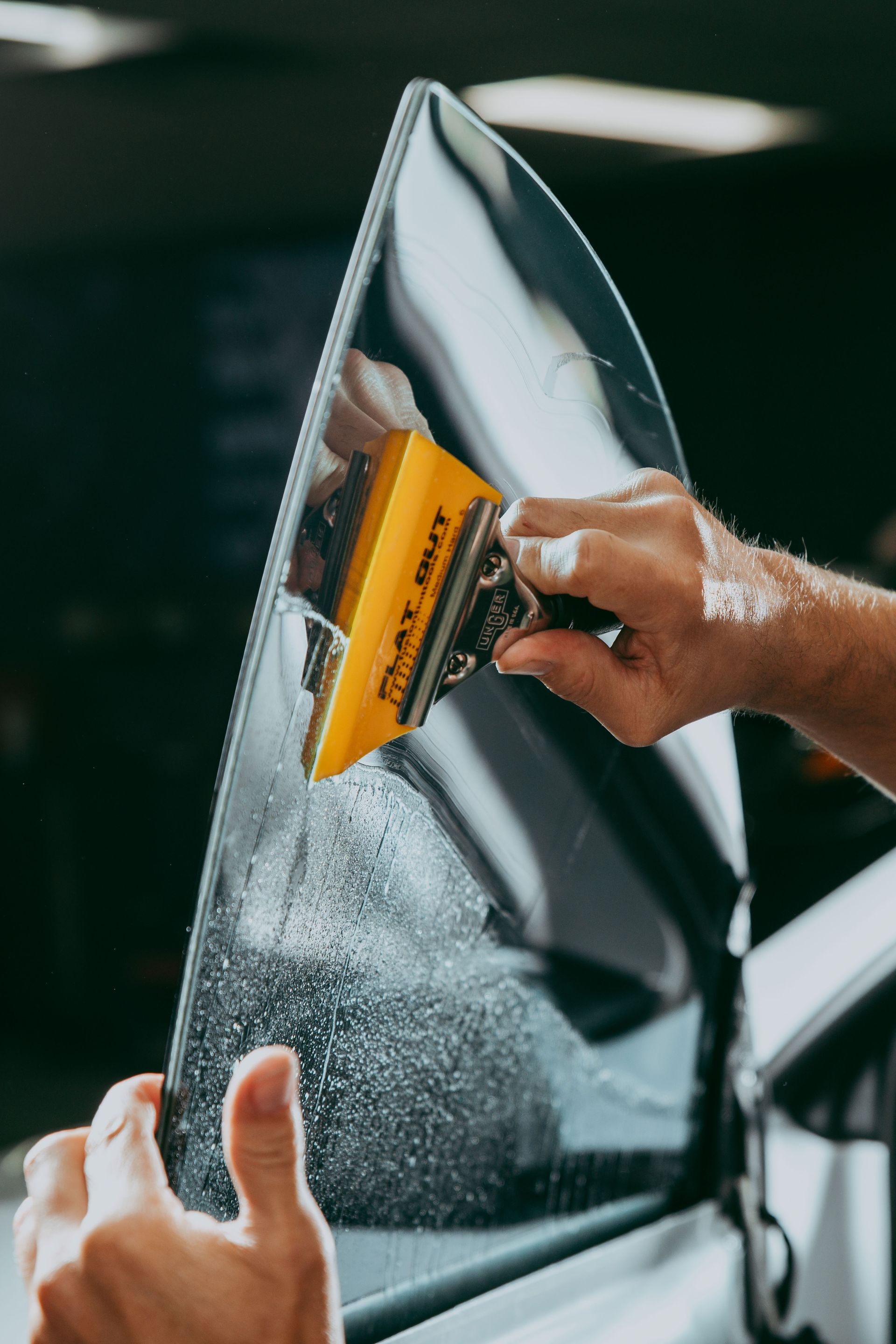 Person applying window tint to a car window with a yellow squeegee.