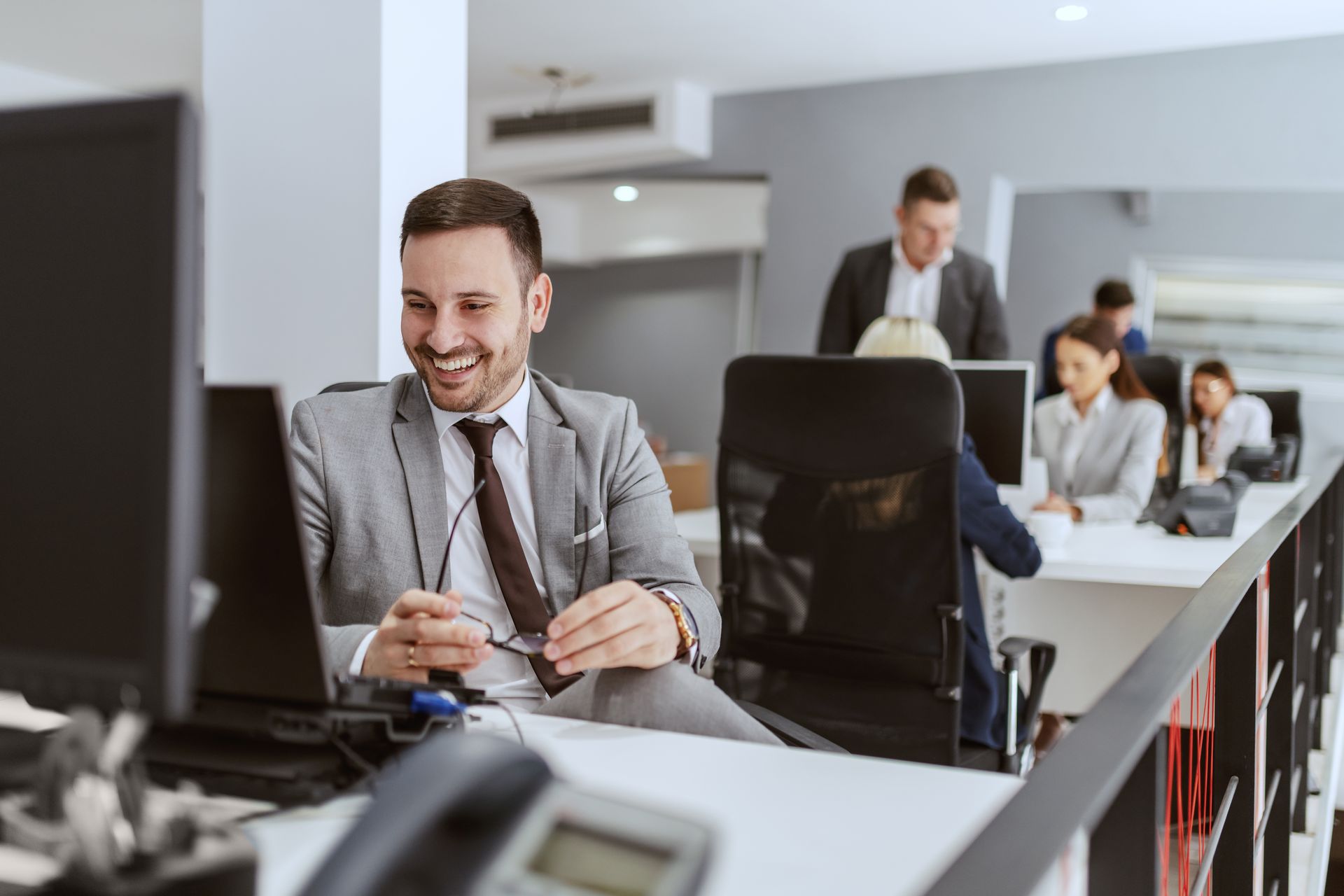 Un hombre con traje gris sonríe frente a una computadora portátil en una oficina moderna; sus colegas en el fondo.