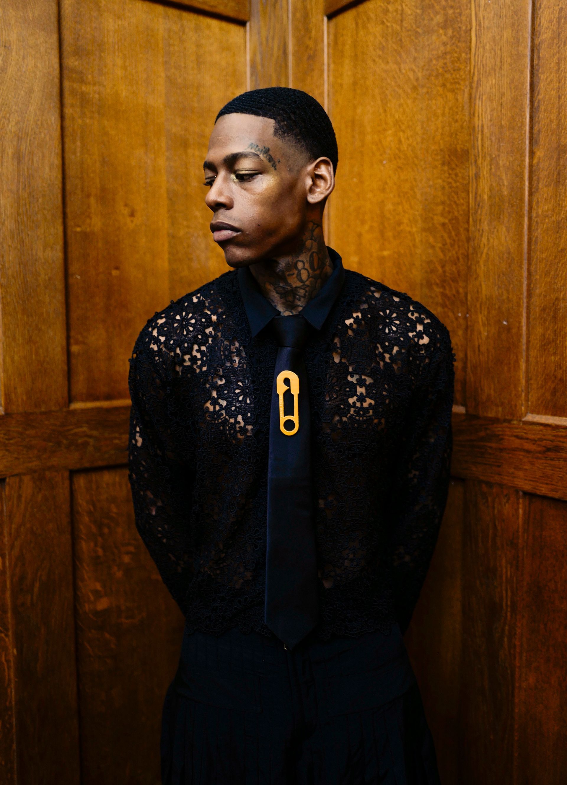 Dash, in dark lace 'Aviary' shirt,  branded tie with safety pin, standing in wood-paneled space, looking away.