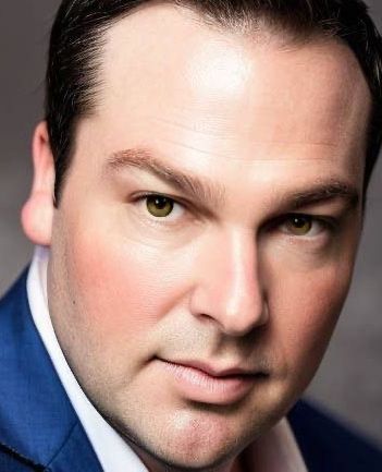 Man with fair skin, wearing a blue jacket and white shirt, looking directly at the viewer.