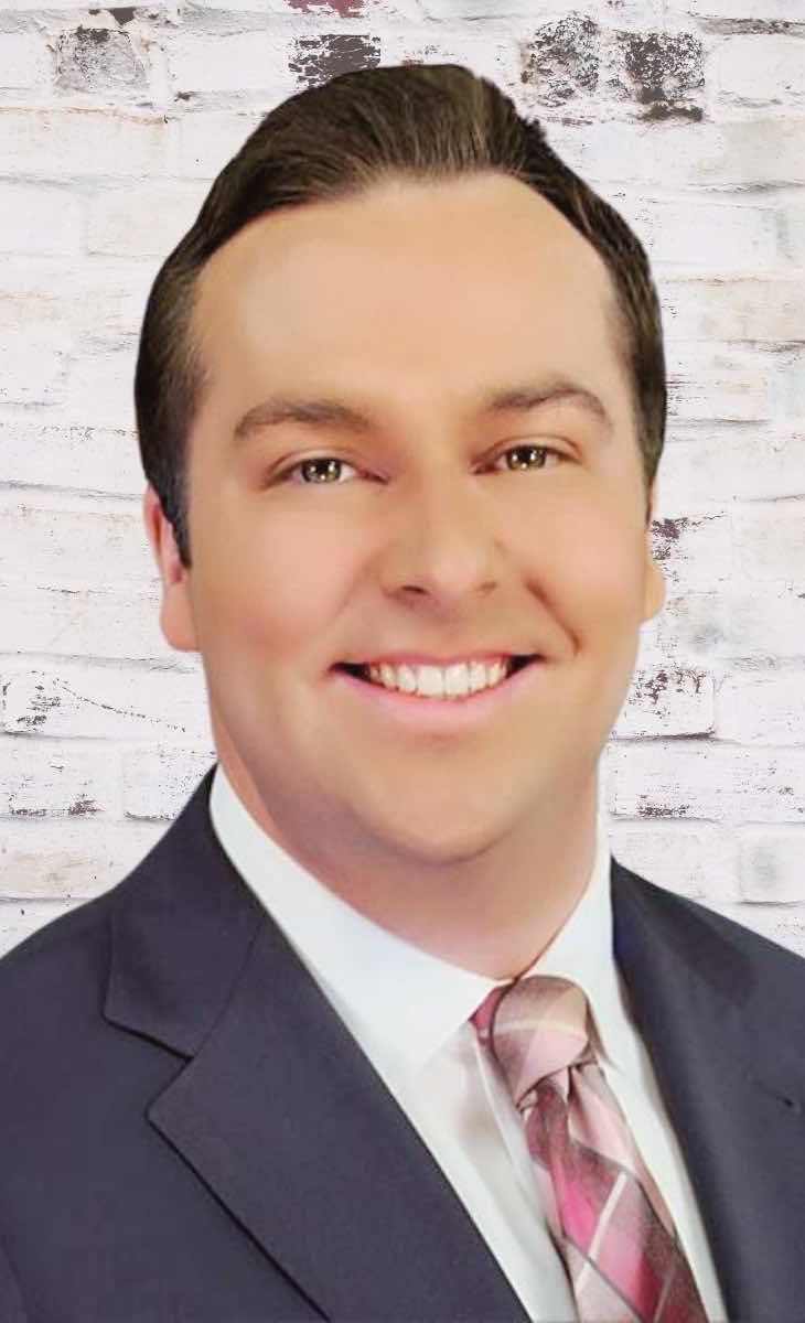 Man in a suit smiles at the camera, with a brick wall background.