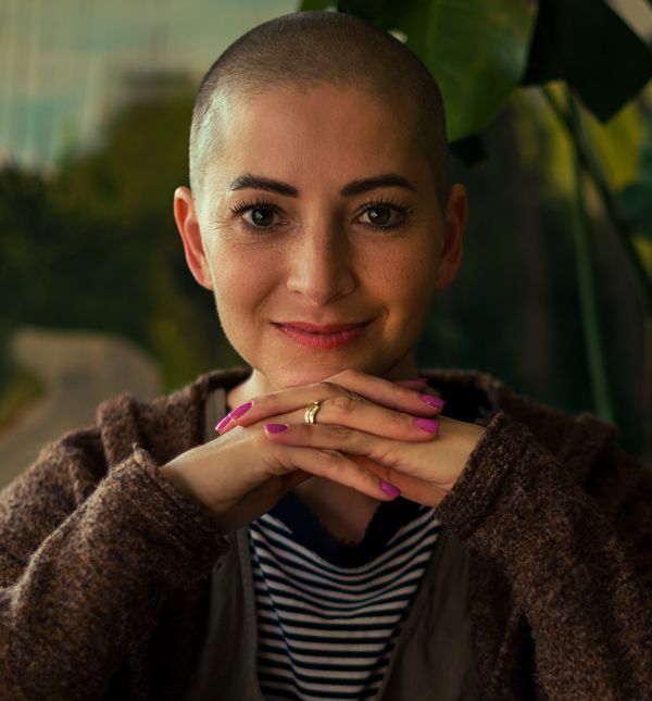 A woman with cancer and a shaved head is wearing a striped shirt and a brown jacket, smiling aftera  free consultation with NYC Acupuncturist Marc Bystock