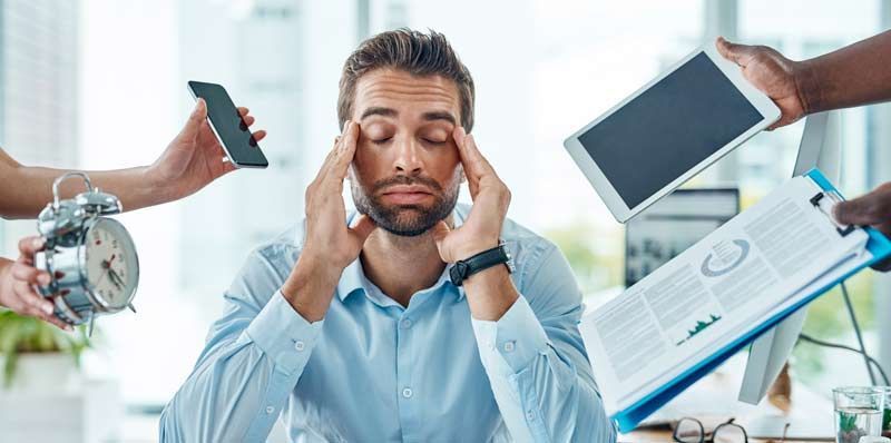 A man stressed at his desk with hands pressing his temples, surrounded by hands holding a clock, phone, tablet, and paperwork need ing Acupuncture for Stress Relief