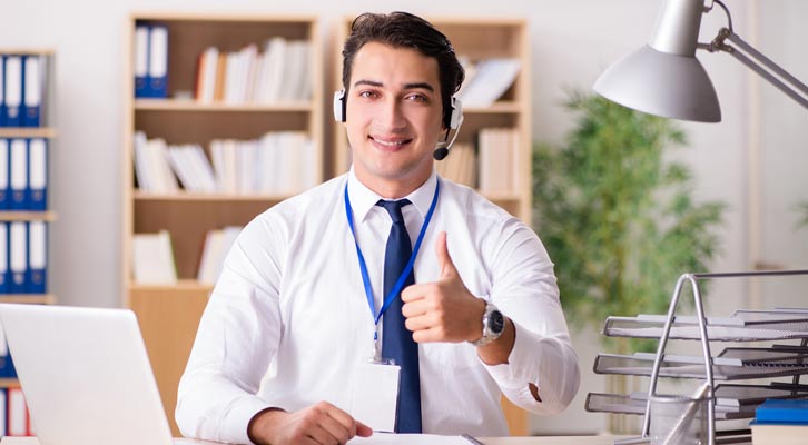 Man wearing headset at desk gives thumbs up in office.