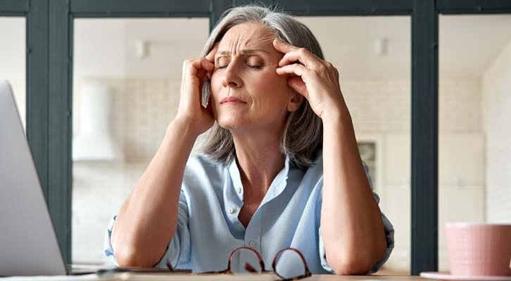 A woman with gray hair holds her temples, eyes closed, looking stressed, with a laptop in front of her, calling for Chronic Headache Treatments NYC by Marc Bystock L.Ac.