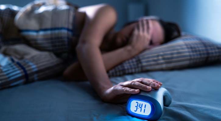 Person in bed reaching to turn off alarm clock, it displays 3:41 AM. Dark room with muted colors. Insomnia Treatments NYC by Marc Bystock L.Ac.