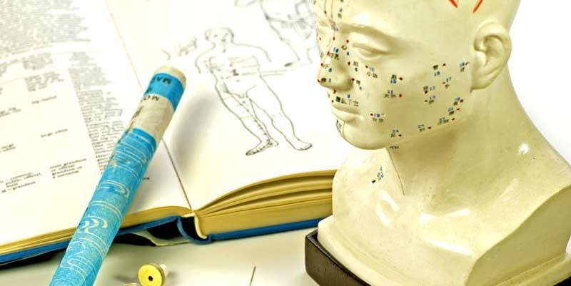 A bust of a human head and open book, marked with acupuncture points. A blue cylindrical object rests on the book representing Integrating Acupuncture with Traditional Medicine: Enhancing Health Through Complementary Practices