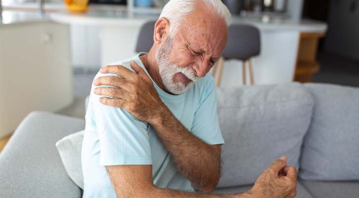 An older man sitting on a couch clutches his right shoulder in pain; indoors, with a light blue shirt and gray beard, needing Shoulder Pain Treatment NYC by Marc Bystock L.Ac.