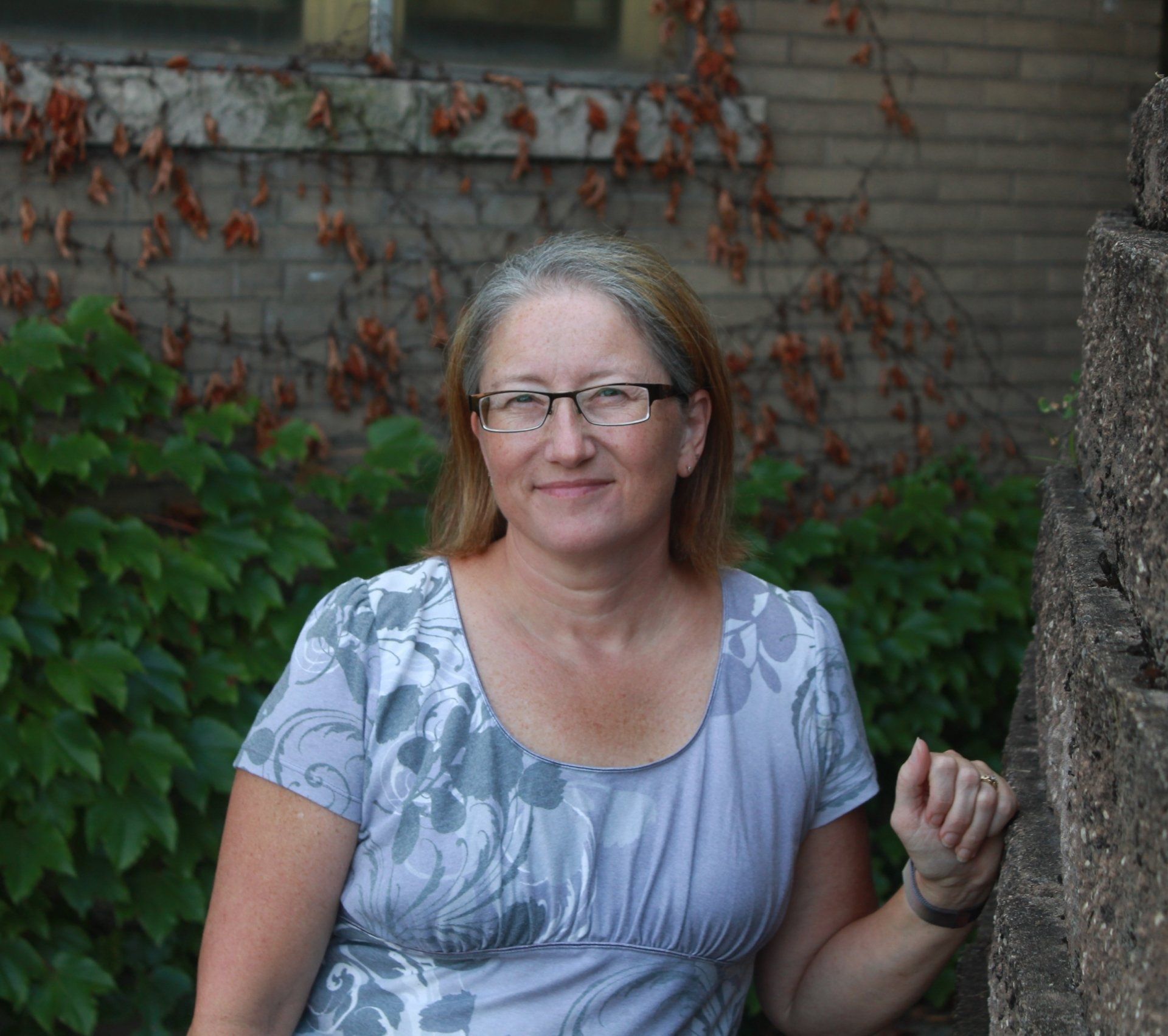 A woman wearing glasses is sitting in front of a brick wall.