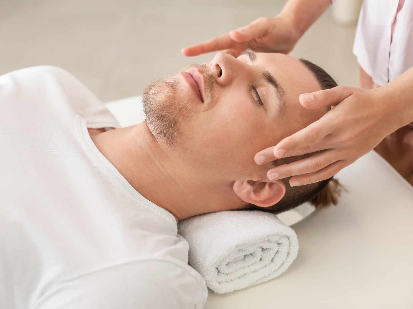 Man receiving head massage, eyes closed, white shirt, light setting.