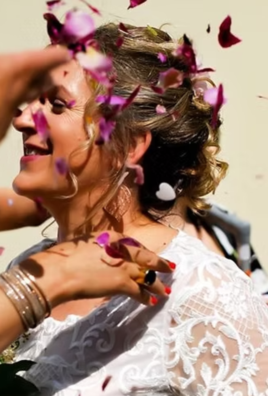 Woman in white lace dress, smiling as flower petals are thrown over her.