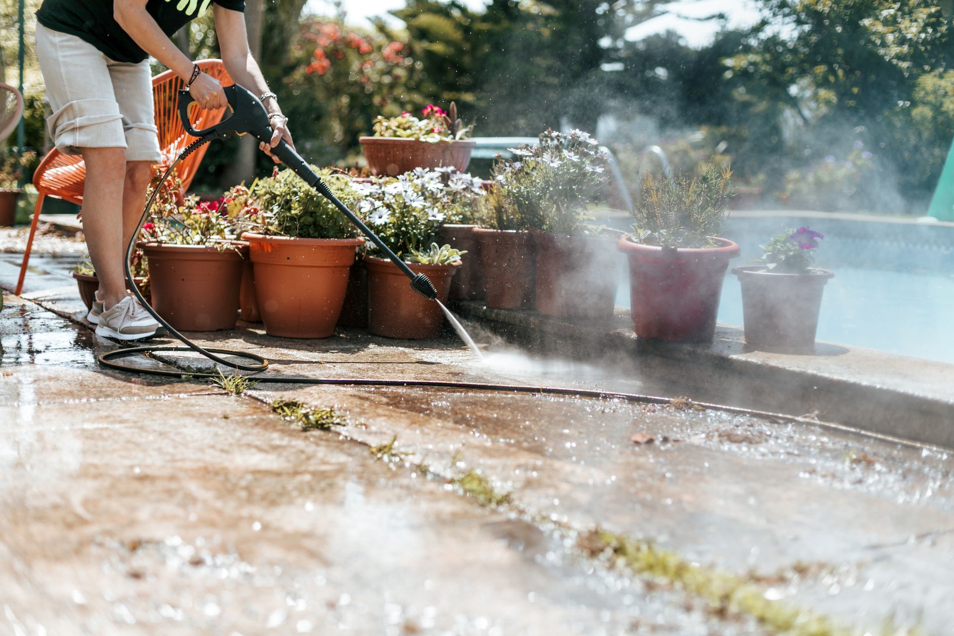 Person using a power washer to clean a concrete patio near a pool and flower pots.
