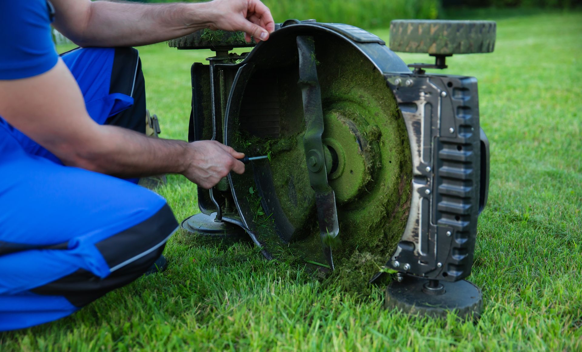 A person in blue overalls cleaning a lawnmower blade in a grassy yard.