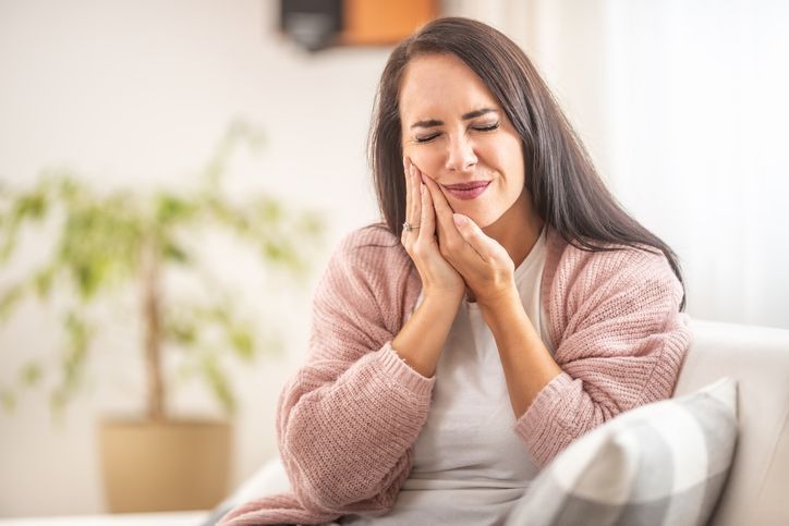 A person in a pink cardigan sits on a sofa, eyes closed, holding their hand to their cheek, appearing to be in pain.