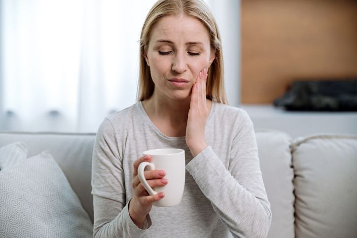 A person sitting on a sofa, holding a white mug and holding their hand to their cheek with a pained expression.