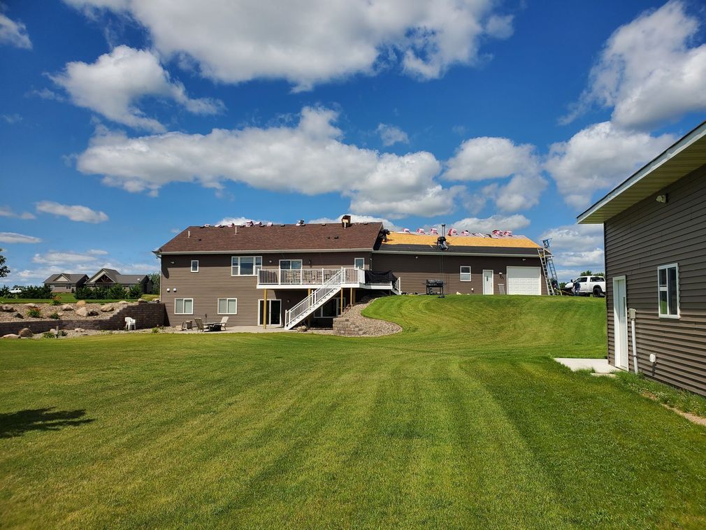 A brown residential house with a partial roof replacement, surrounded by a large green lawn under a sunny, cloudy sky.