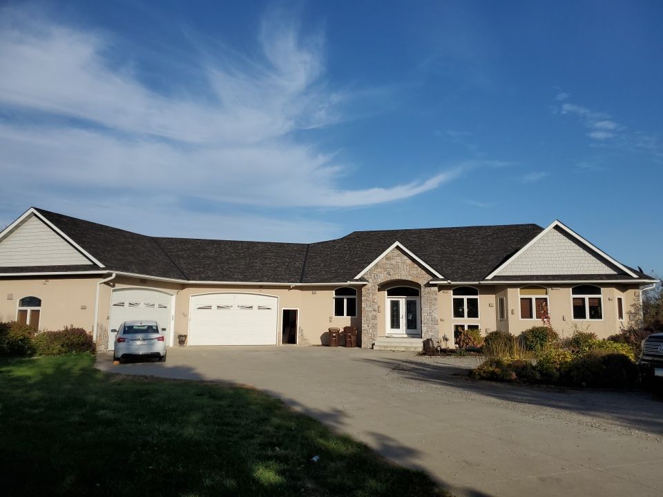 A single-story, beige stucco house with a dark roof and a multi-car garage, seen from a paved driveway under a blue sky.