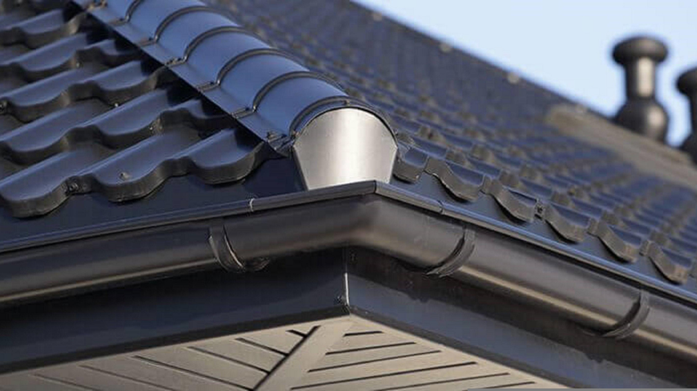 A close-up of a modern dark-tiled roof with a metal ridge cap, a gutter system, and an eaves soffit.