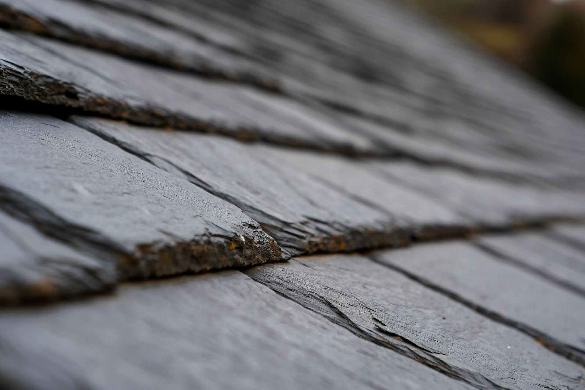 A person in work pants stands on a residential roof, pulling up a section of damaged, layered asphalt shingles.