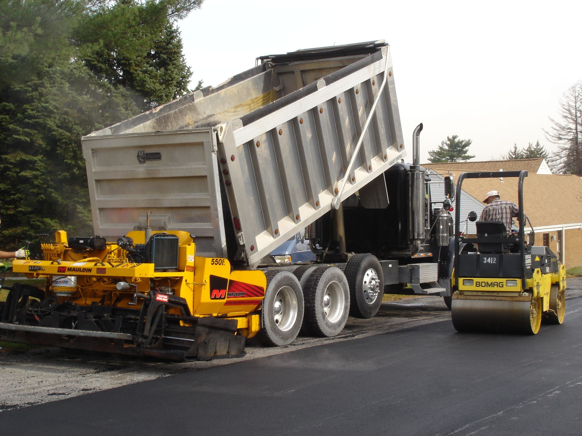 A dump truck unloads materials into a yellow asphalt paver next to a road roller on a construction site.