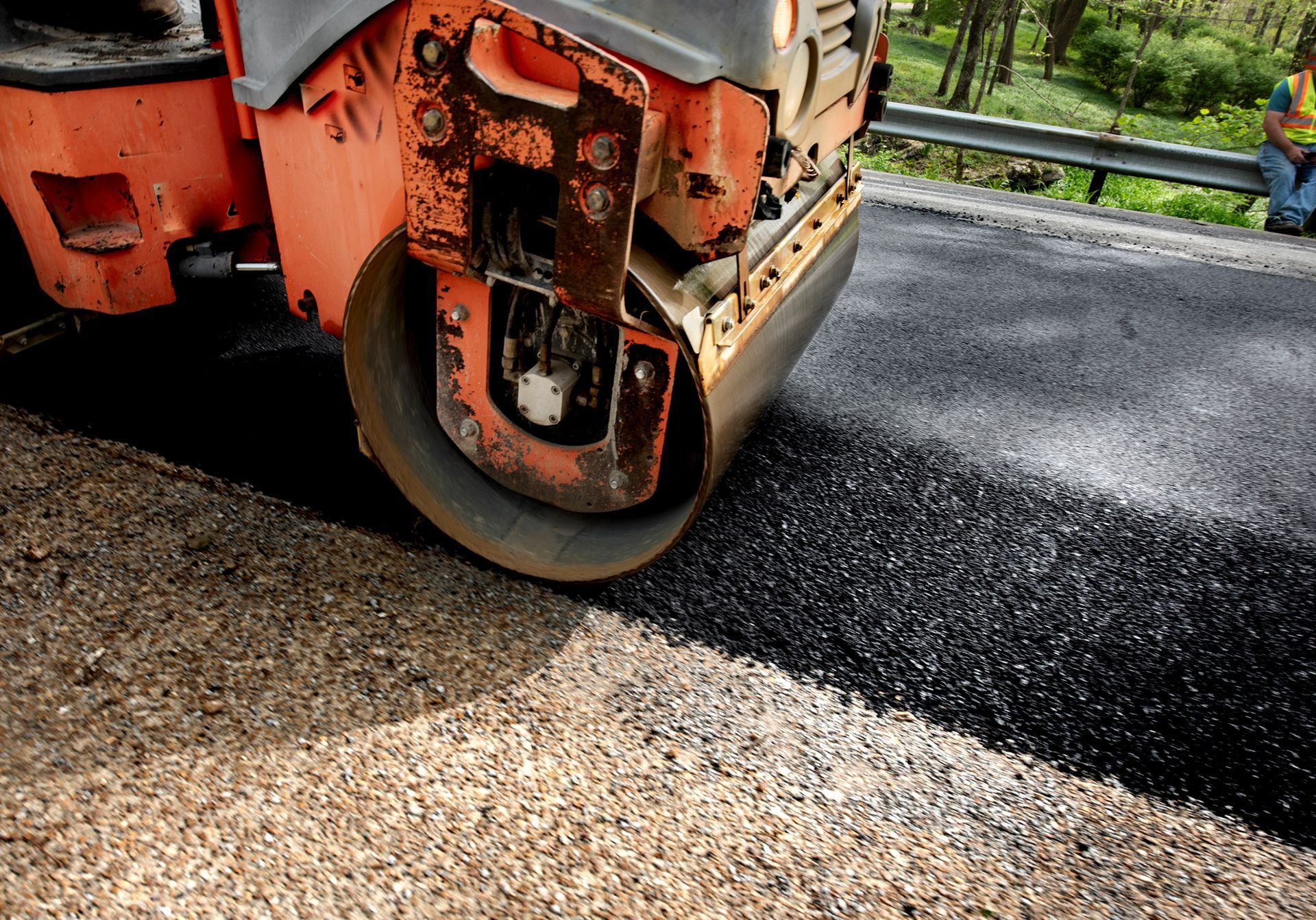 Road roller compacting fresh asphalt on a roadway; orange machine, black surface, rural setting.