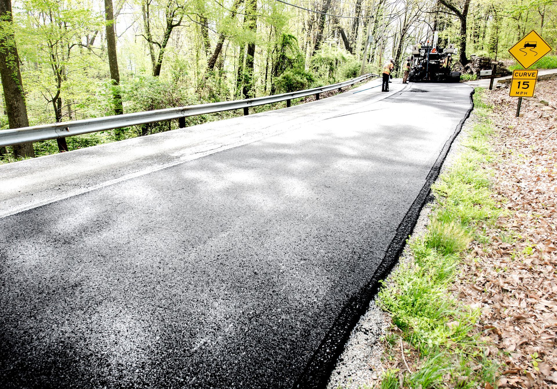 Freshly paved asphalt road next to a metal guardrail and grassy verge in a wooded area.