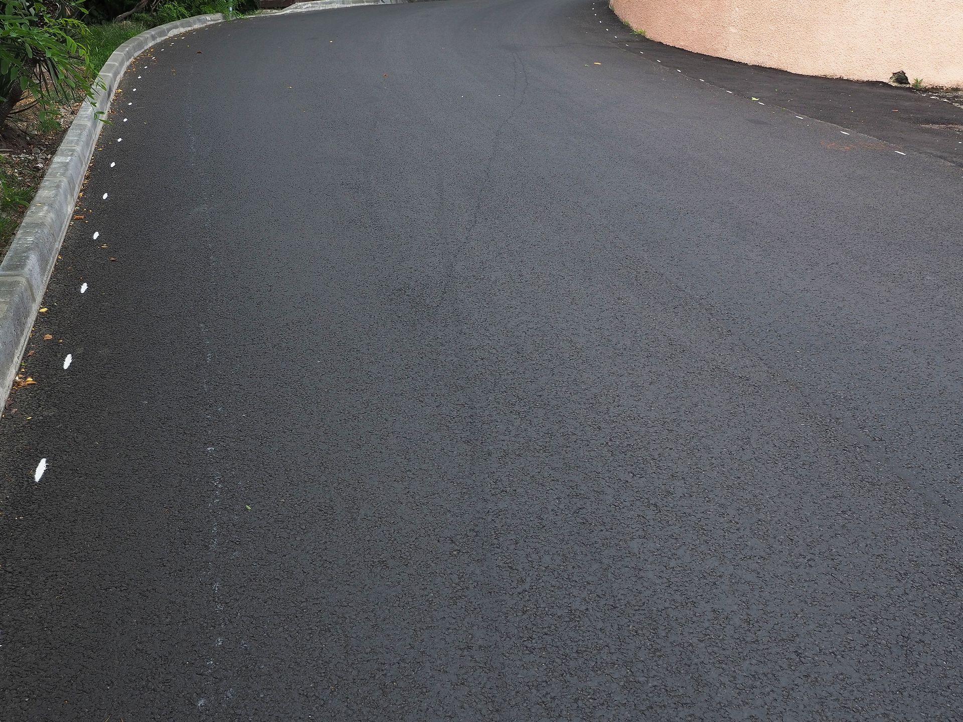 Black asphalt road curving towards a tan wall, with white markers along the curb.