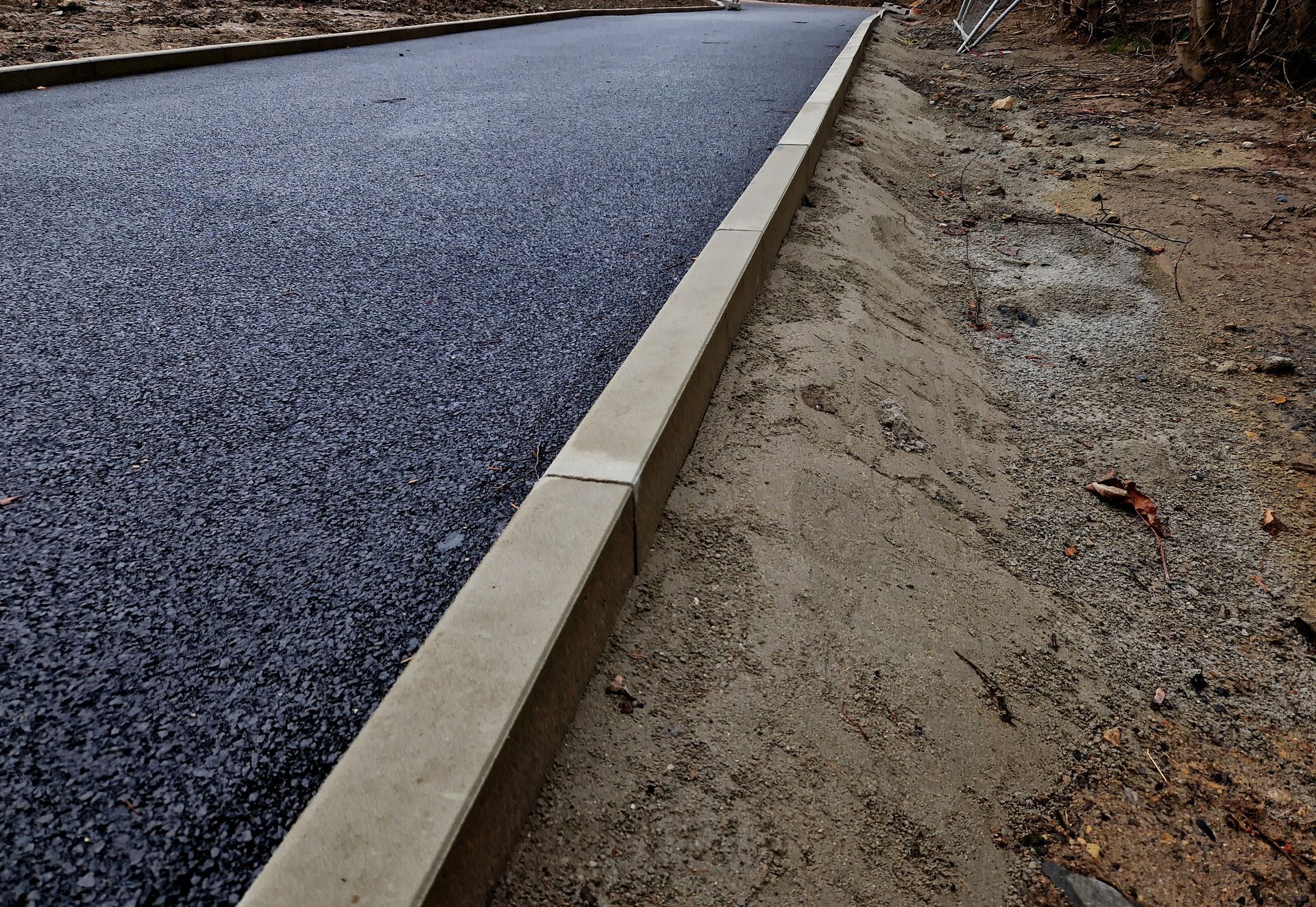 Newly paved asphalt road with concrete curb bordering dirt.