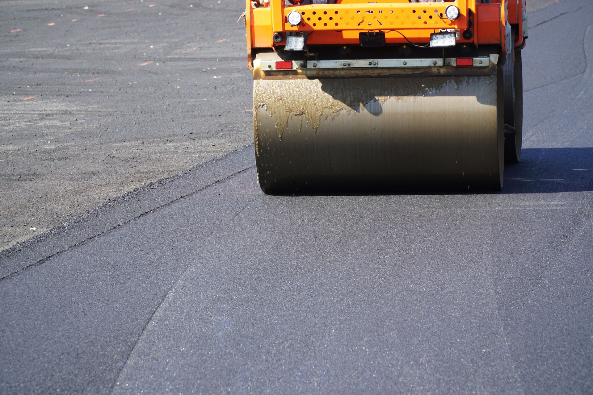 Road roller compacting fresh asphalt; orange machine, gray drum.