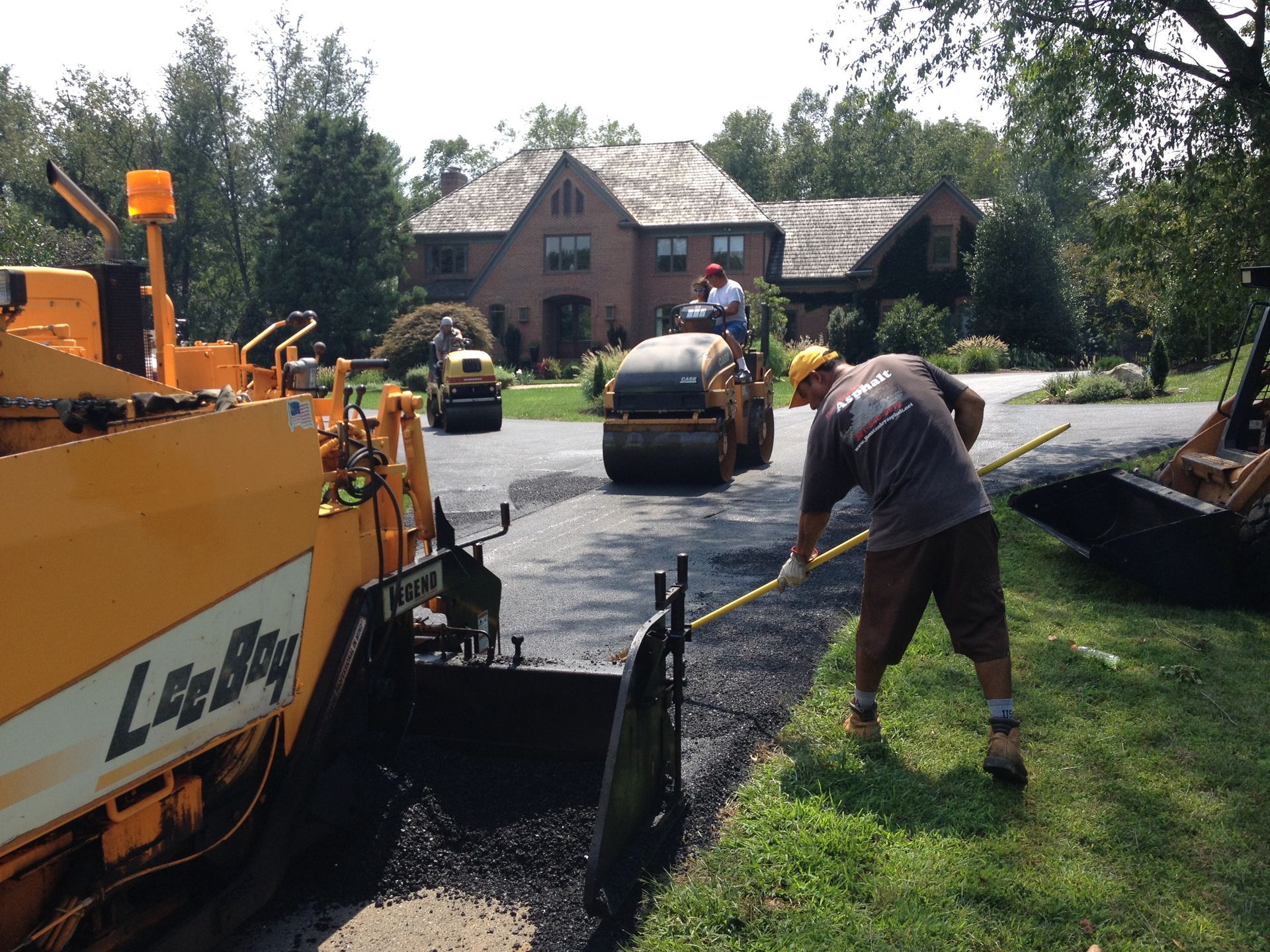 Workers use heavy machinery to pave a driveway with asphalt in front of a brick house.