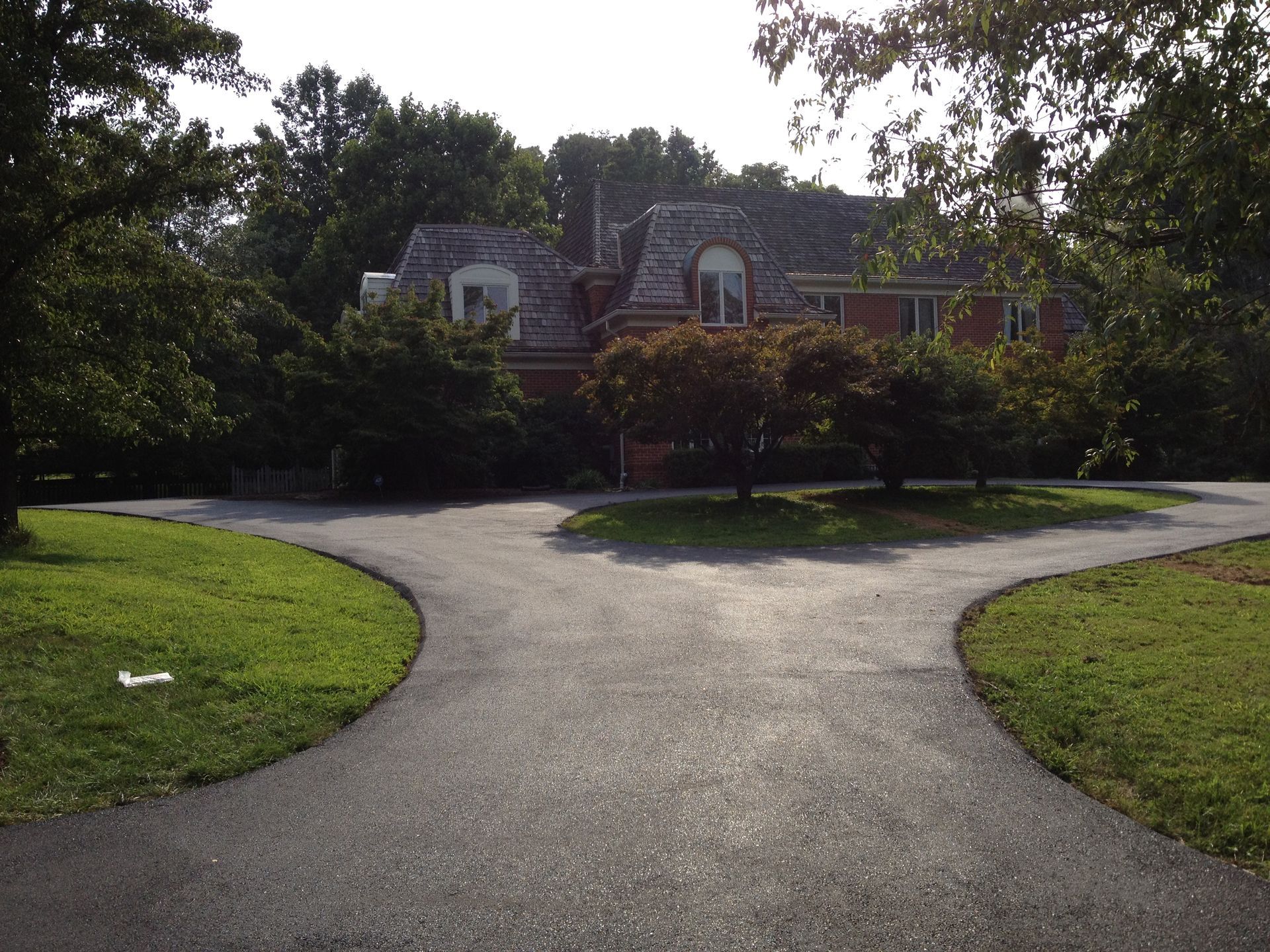A large red brick house sits at the end of a Y-shaped paved driveway, surrounded by green lawns and mature trees.