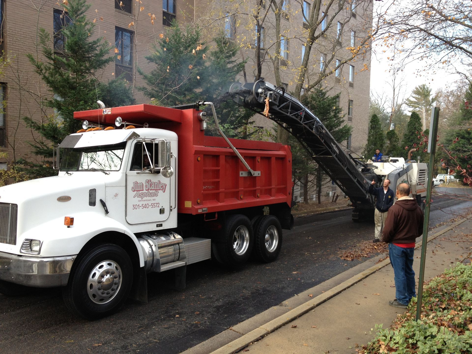 A red dump truck parked on a street with a mechanical arm loading leaves into the back, with two workers standing nearby.