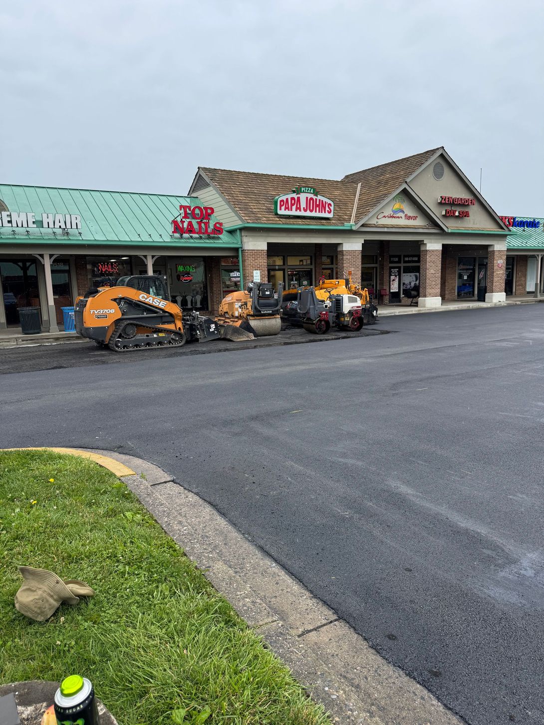 Asphalt paving in progress at a strip mall. Heavy machinery works on a dark, wet surface.