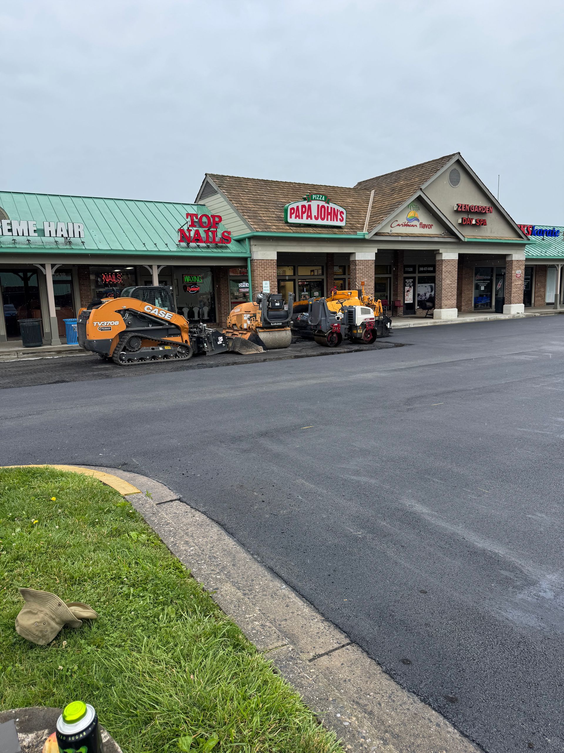Asphalt paving in progress at a strip mall. Heavy machinery works on a dark, wet surface.