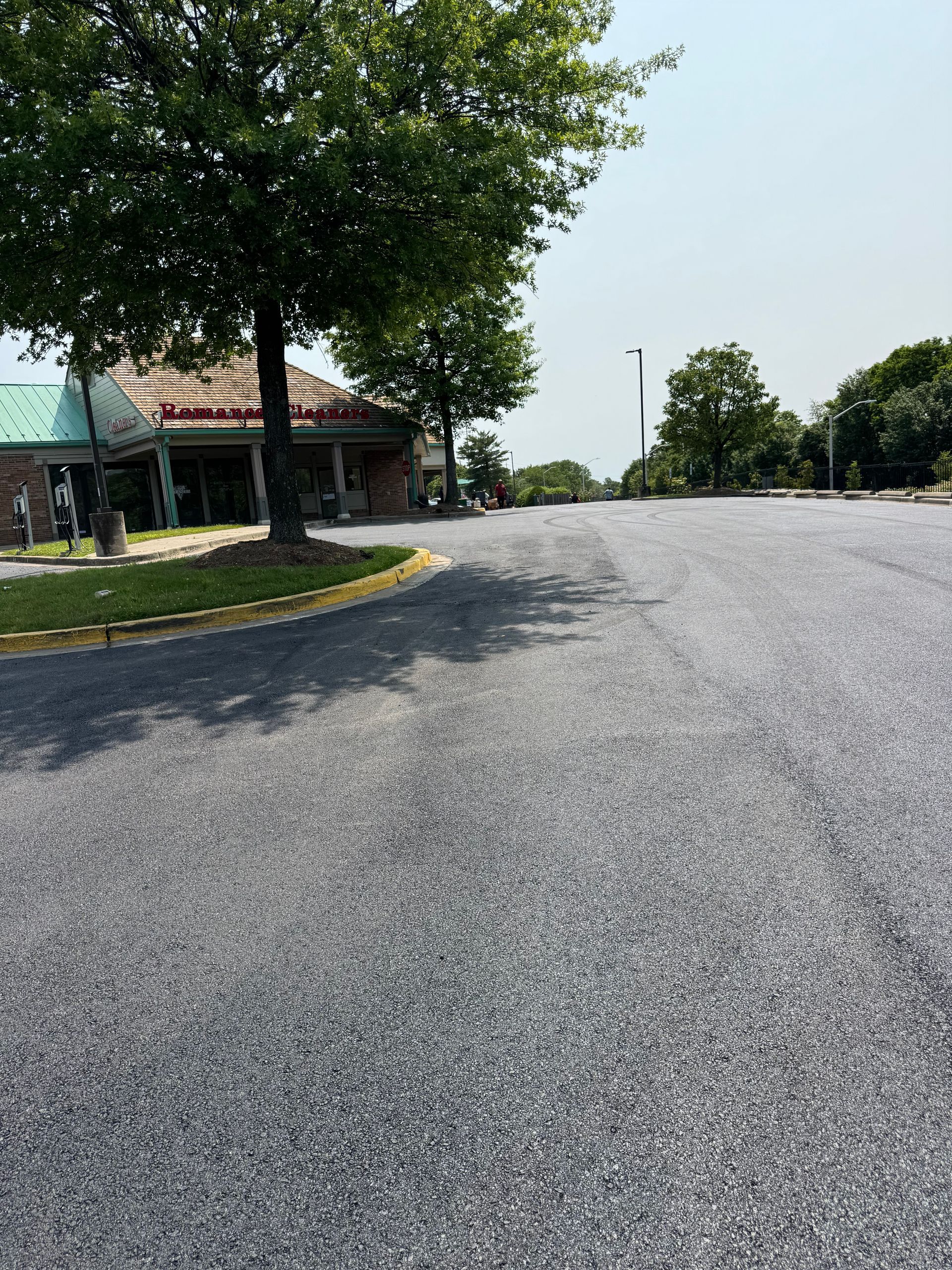 Asphalt road curves past a building with a red tile roof and trees under a sunny sky.