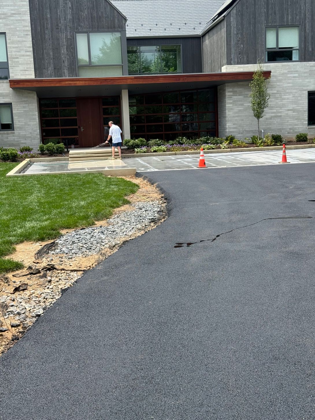 Person sprays water on a driveway in front of a modern building. Asphalt driveway, green lawn, and orange cones visible.